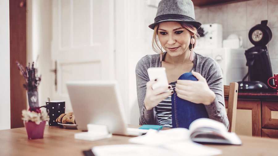 Jonge vrouw aan tafel met haar smartphone, een notitieboekje en documenten om haar huishoudboekje bij te houden.